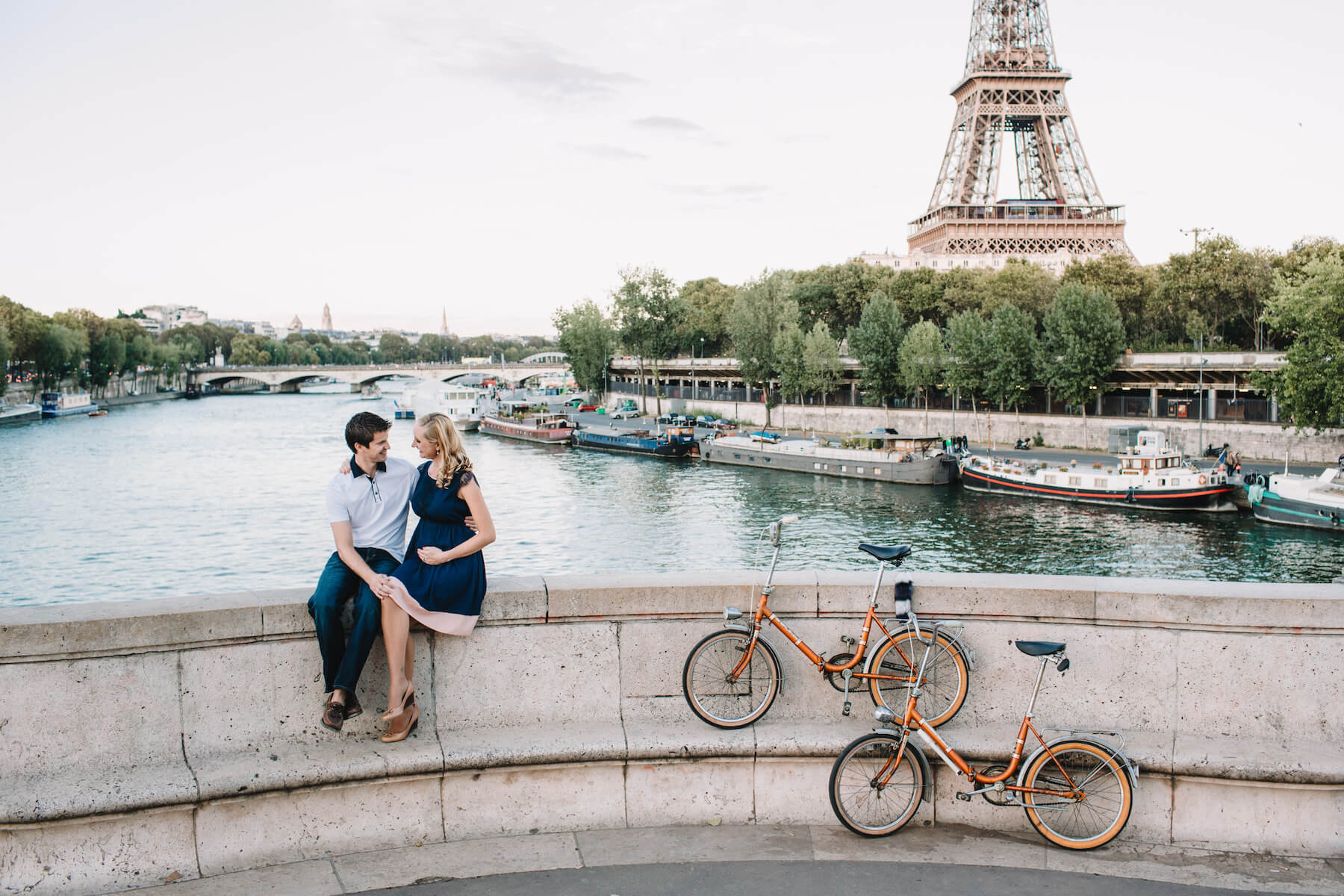 a couple sitting on a ledge, she is pregnant and holding her belly in front of the Eiffel Tower in Paris, France