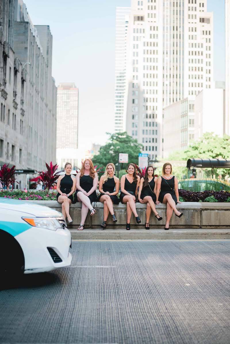 Six female friends on a bachelorette trip together sitting on a stone ledge along a sidewalk in Chicago, USA