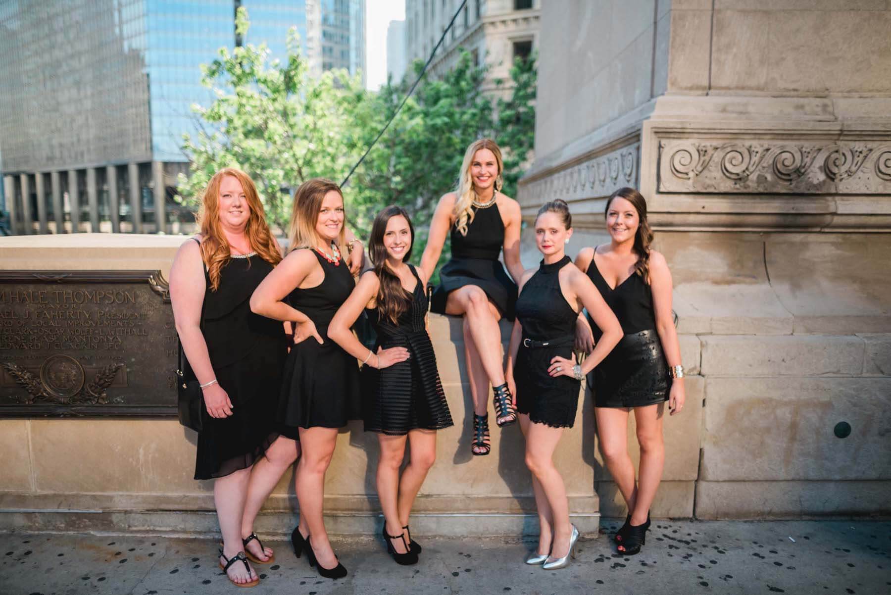 Six female friends standing together with bride-to-be sitting up on a ledge on a bachelorette trip in Chicago, USA