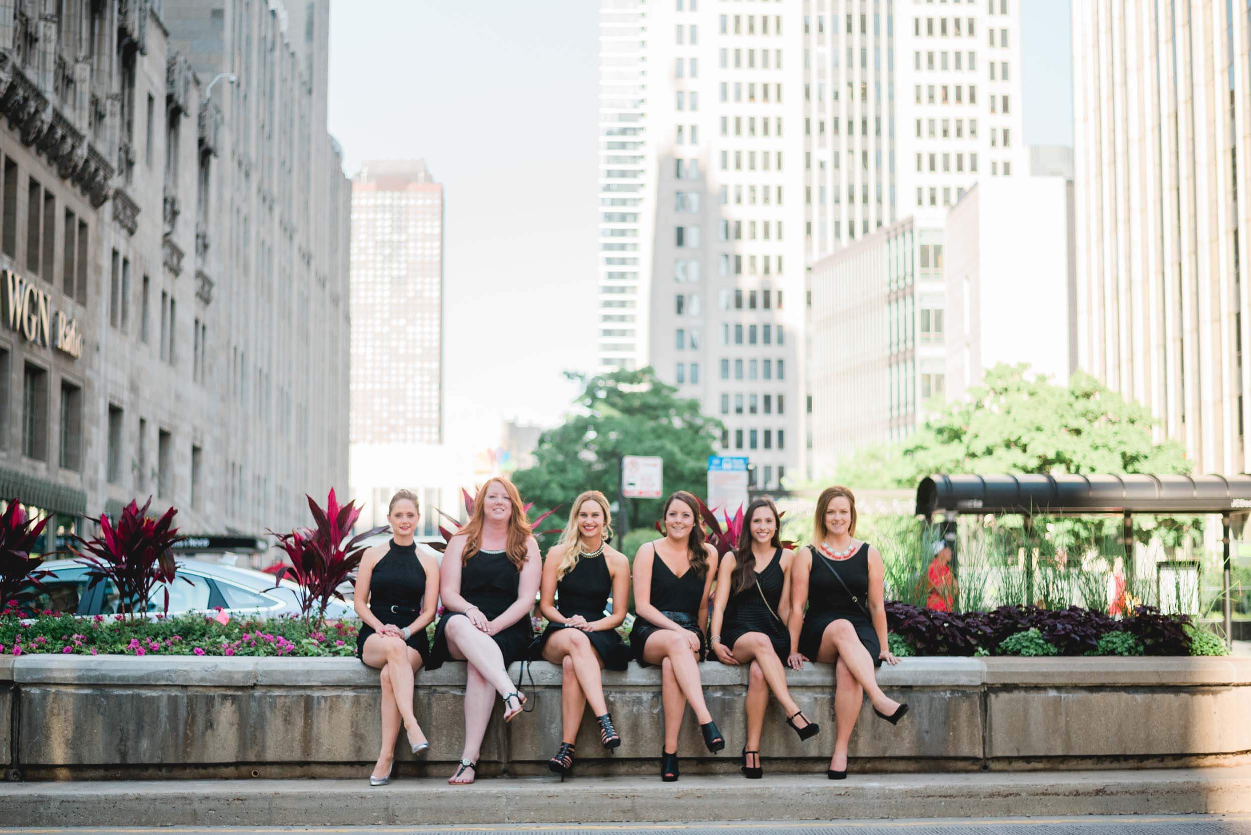 Six female friends sitting together on a stone ledge along a sidewalk on a bachelorette trip in Chicago, USA