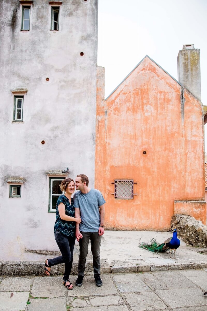 Couple standing close to one another and smiling while standing near a peacock bird on a couples trip in Lisbon, Portugal