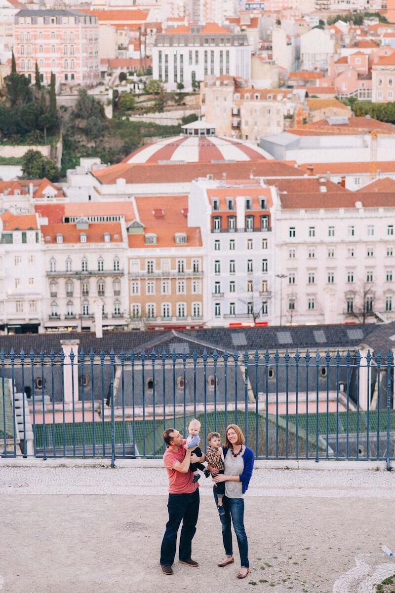 Family of four with two young sons smiling at the Miradouro de São Pedro de Alcântara in Lisbon, Portugal
