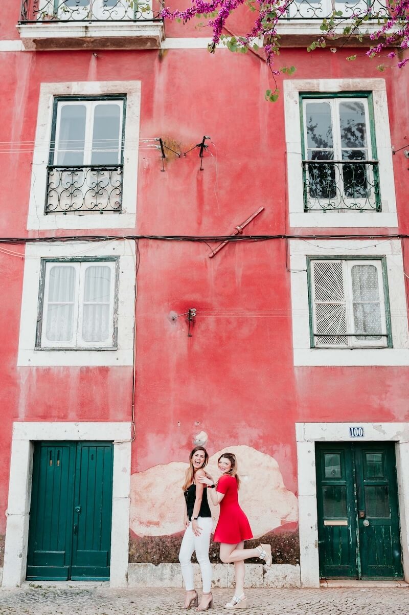 Two female friends standing together in front of a bright red building on a friends trip in Lisbon, Portugal