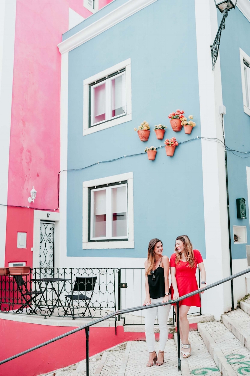 Friends smiling at each other while standing on some steps in the Rua da Galé district of Lisbon, Portugal