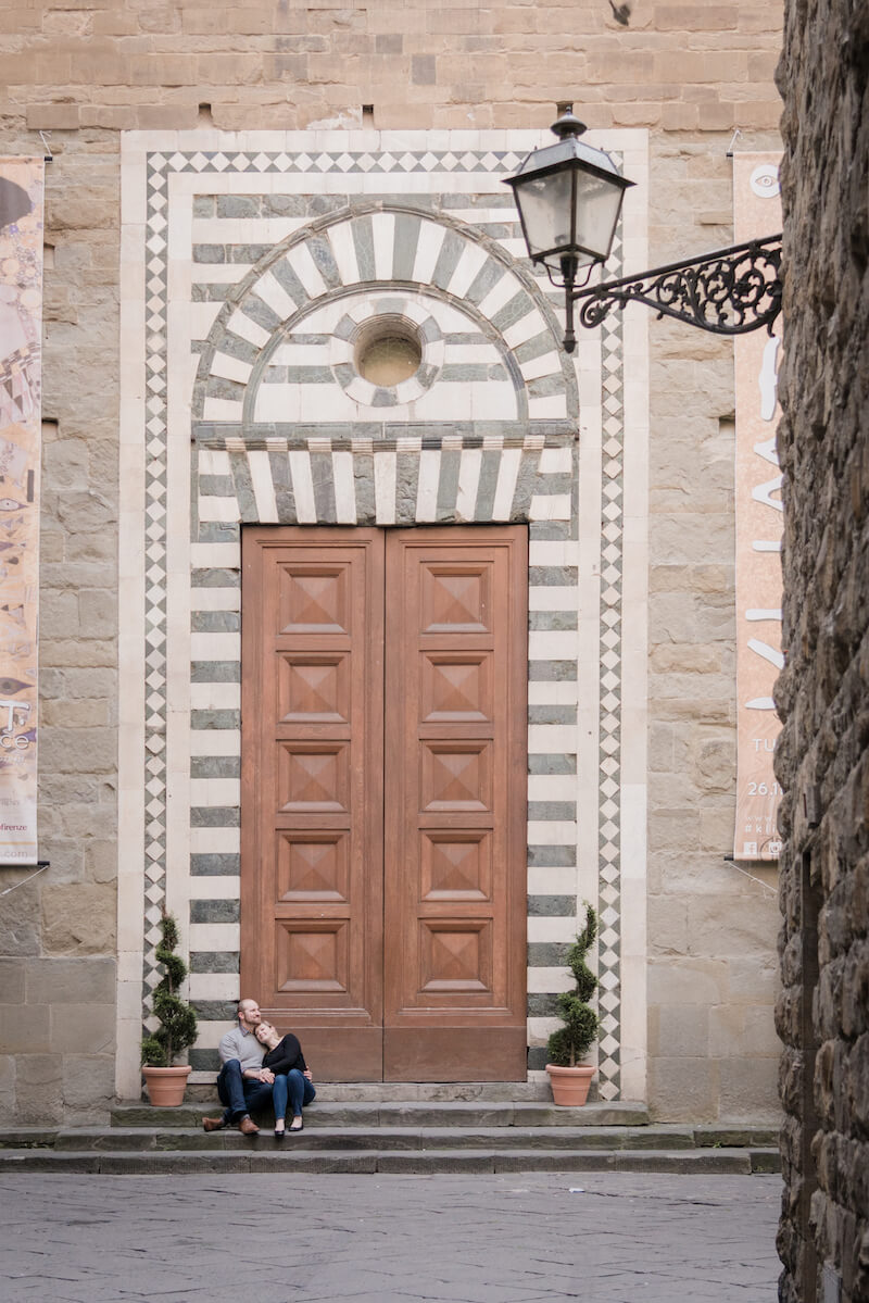 Couple sitting on steps together in front of giant wooden door in Florence, Italy