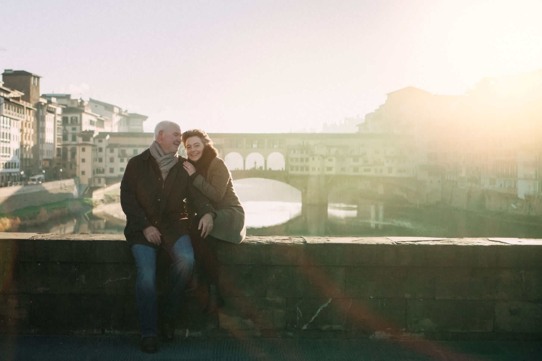 Mature couple sitting together in the winter on bridge ledge in front of Ponte Vecchio in Florence, Italy