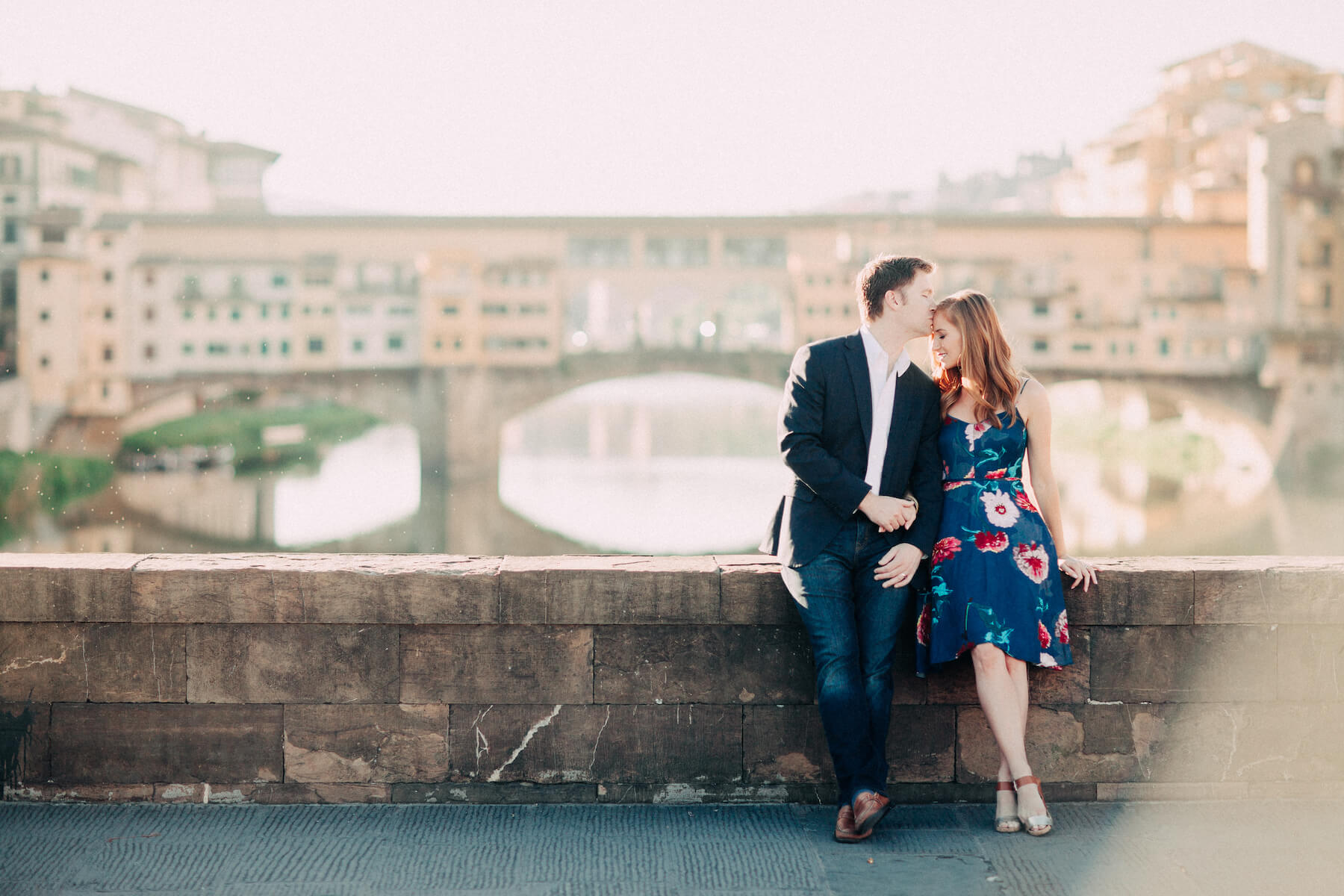 Man kissing his partner's forehead, sitting on bridge ledge in front of Ponte Vecchio on couples trip in Florence, Italy