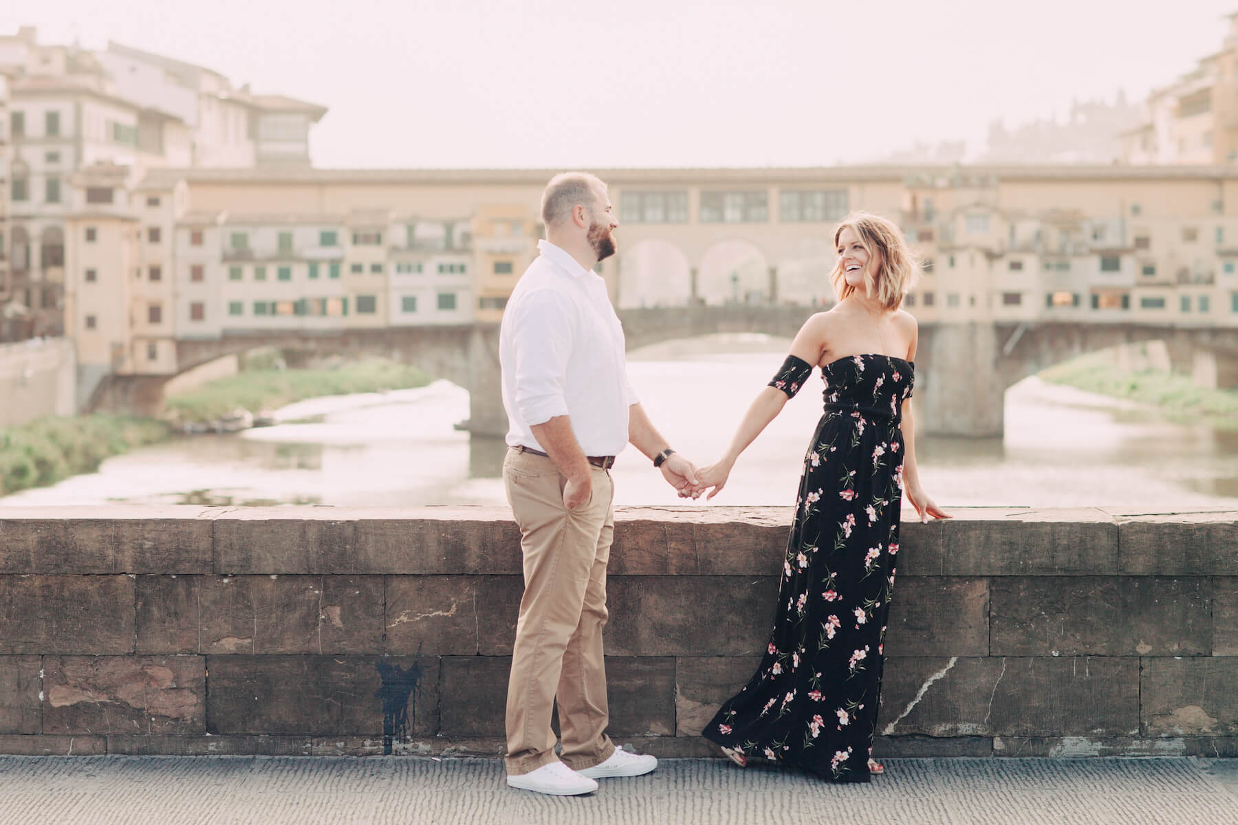 Couple standing on bridge holding hands in front of Ponte Vecchio on couples trip in Florence, Italy