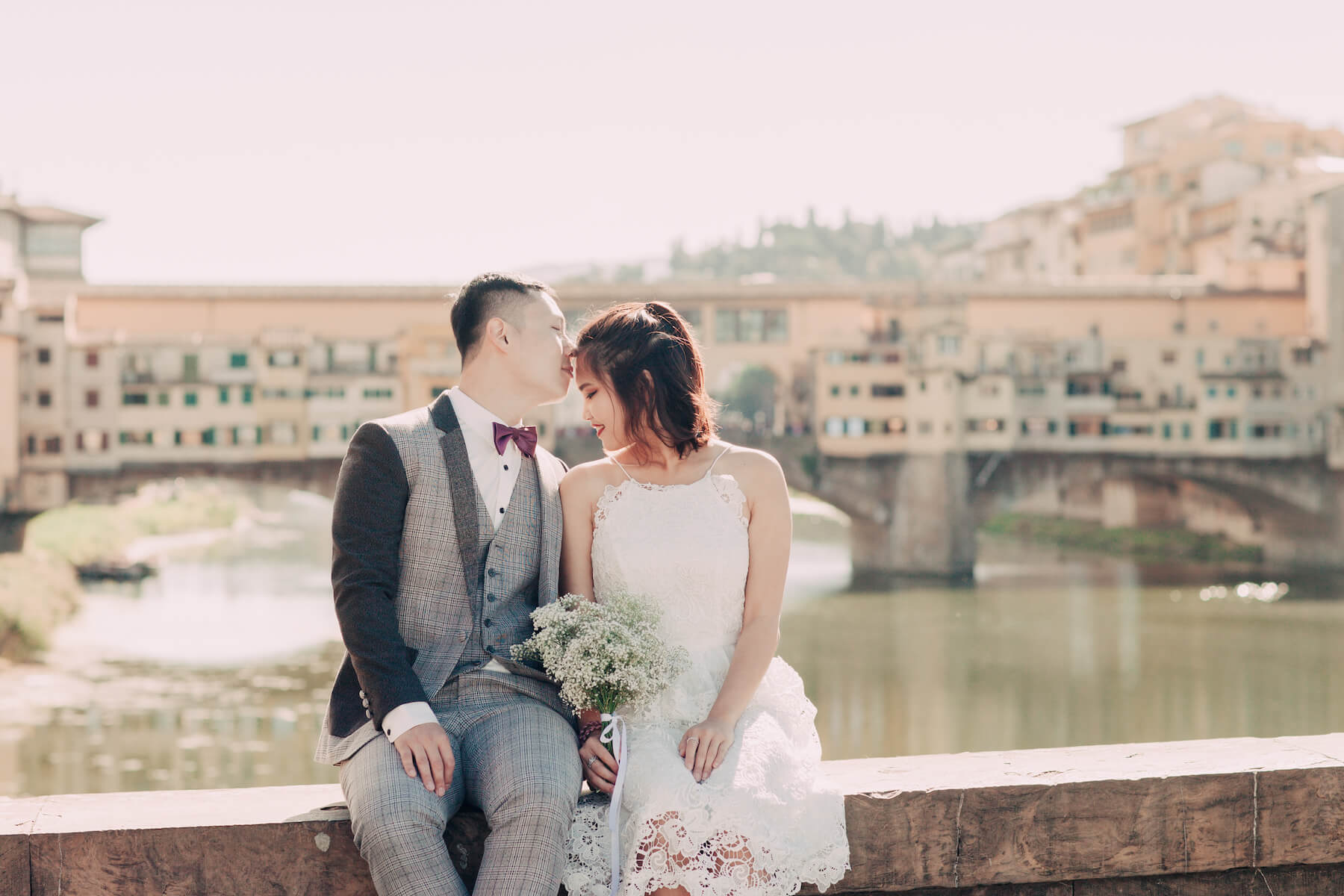 Man kissing partner's forehead sitting on bridge ledge together at Ponte Vecchio in Florence, Italy