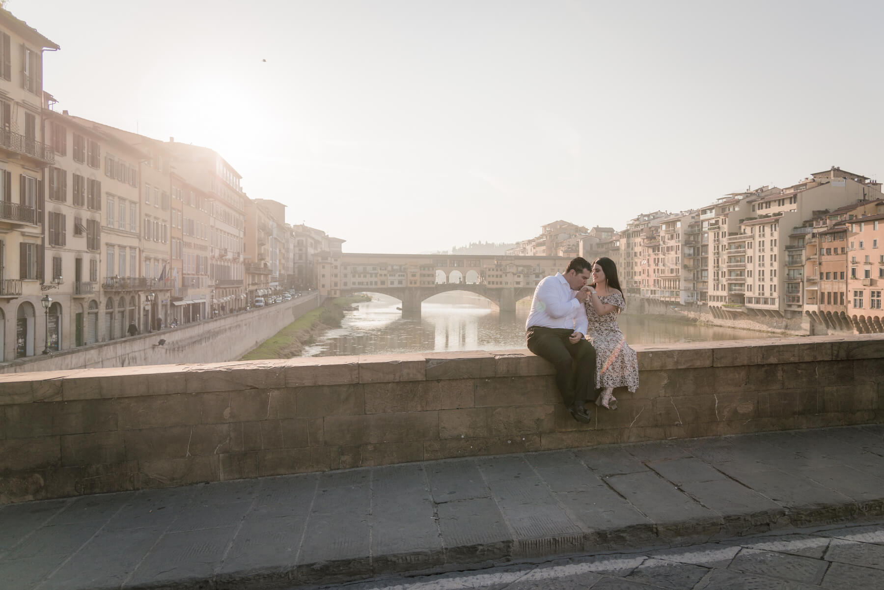 Man kissing partner's hand sitting on bridge ledge together at Ponte Vecchio in Florence, Italy