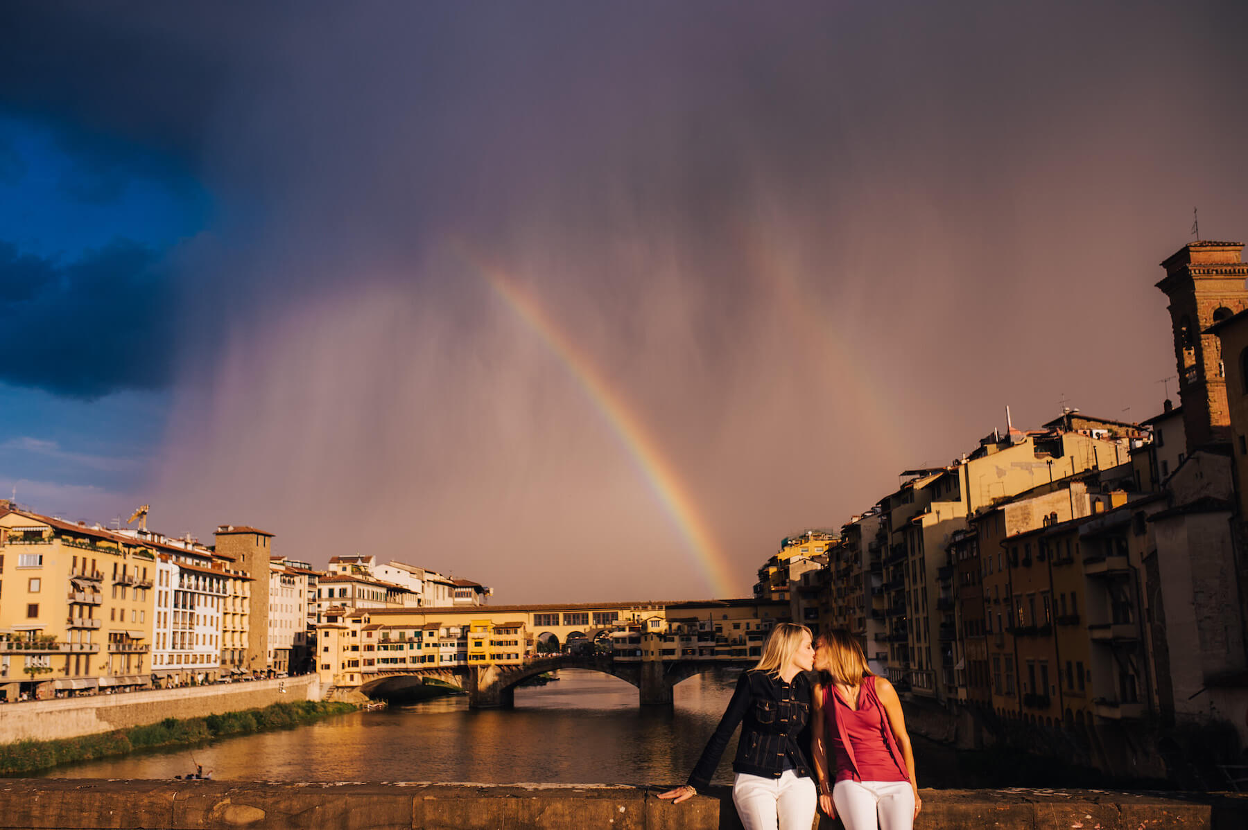 LGBTQ+ couple kissing while sitting on a bridge ledge together with a rainbow in the background in Florence, Italy