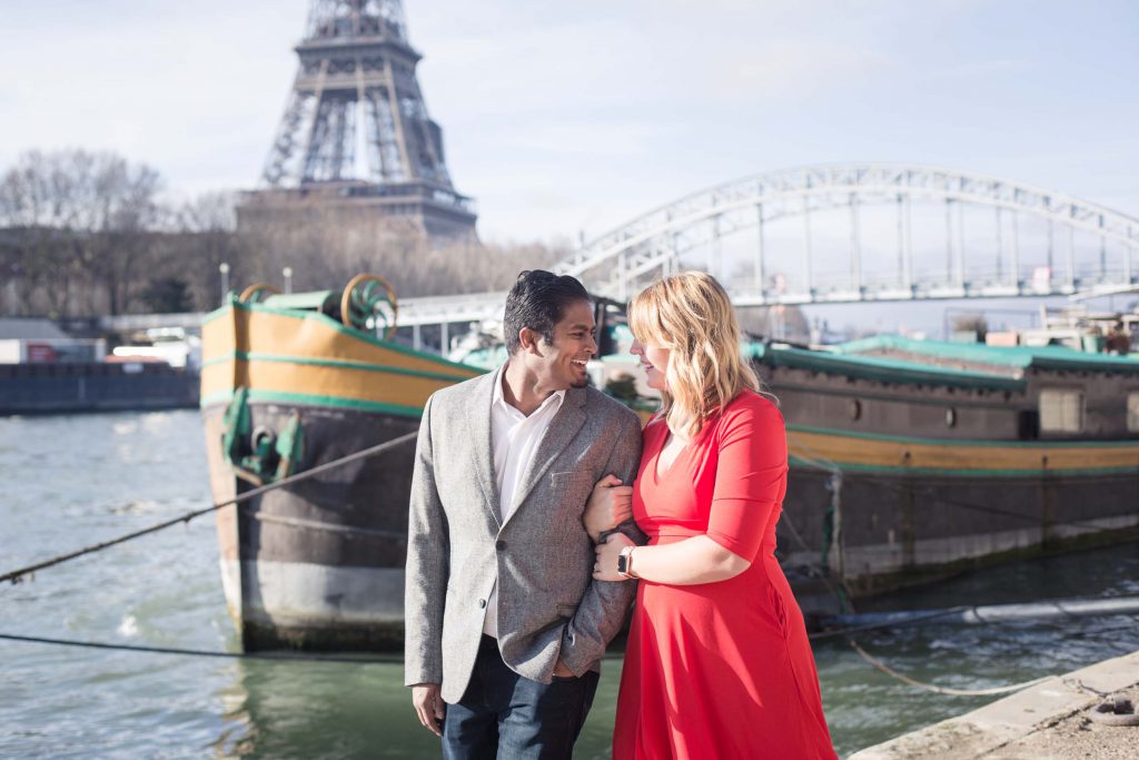 A couple walks arm in arm along the Seine in Paris with the Eiffel Tower in the background.