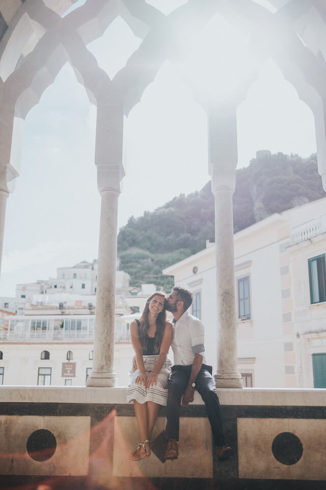Couple sitting on a wall ledge beneath an archway with the male kissing his female partner's forehead in Amalfi Coast, Italy