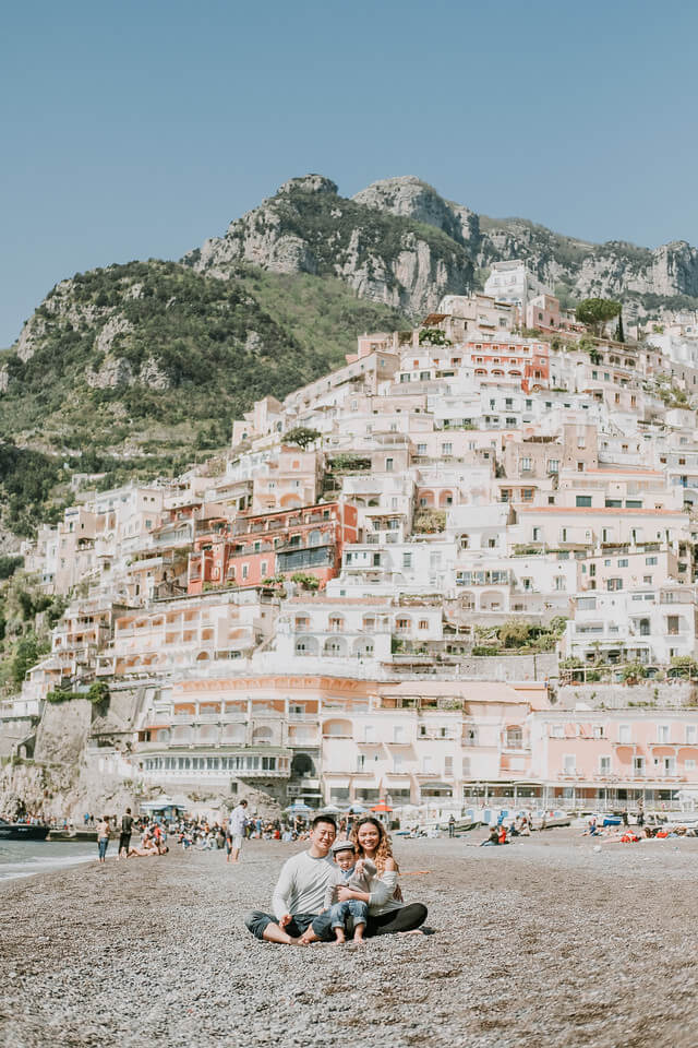 Family sitting on the beach together with the Amalfi Coast pastel pink buildings in the background in Italy