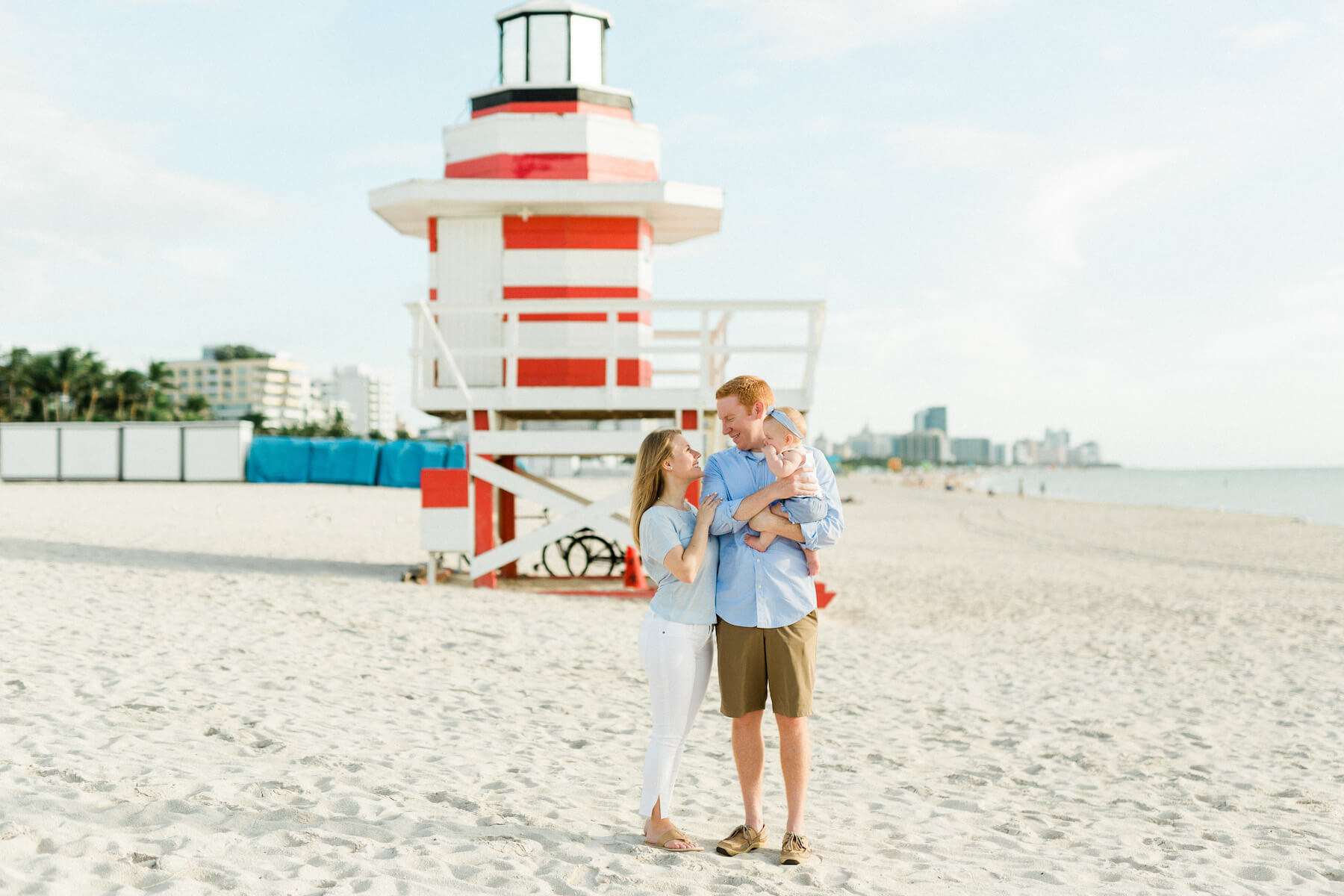 A family with a baby standing on the beach, in front of a colourful lifeguard station in Miami, Florida