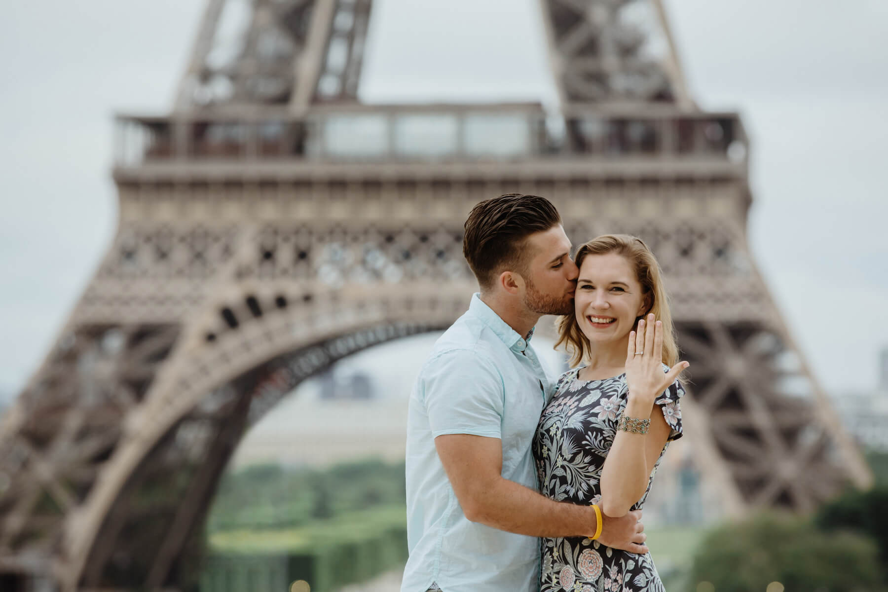couple in front of the Eiffel Tower, he just proposed and she is holding holding her hand up in Paris France
