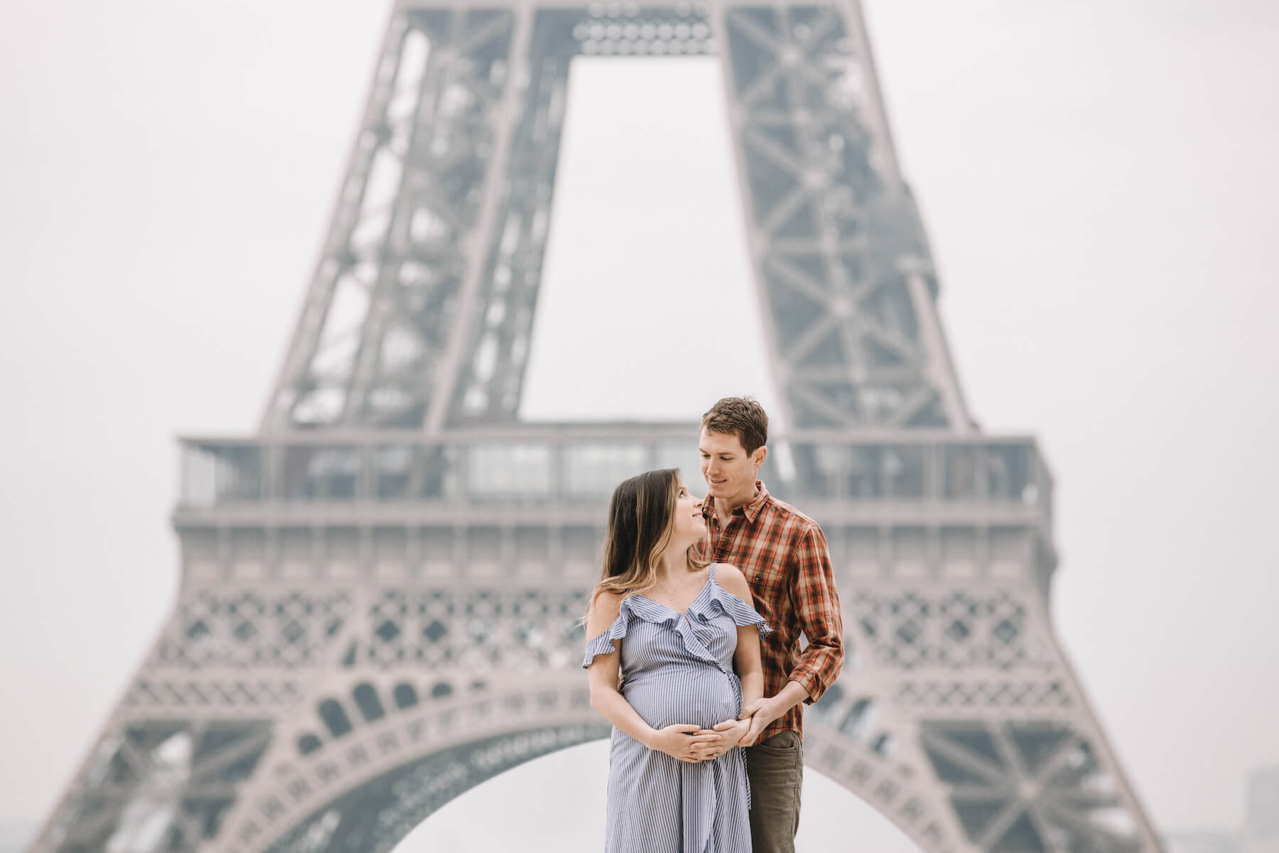 couple in Paris standing in front of the Eiffel tower, the woman is pregnant and they are holding her stomach in Paris, France