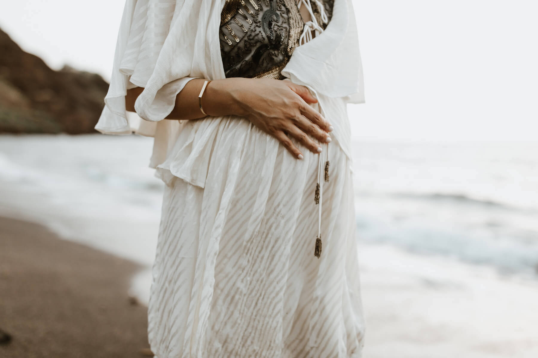 pregnant woman holding her stomach on the beach, wearing beautiful white dress in Maui, Hawaii