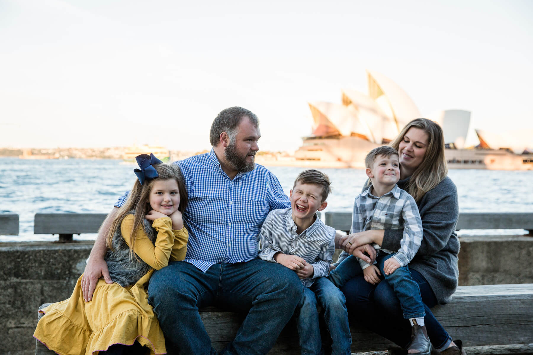 a family of four sitting down with the Opera House behind them in Sydney, Australia
