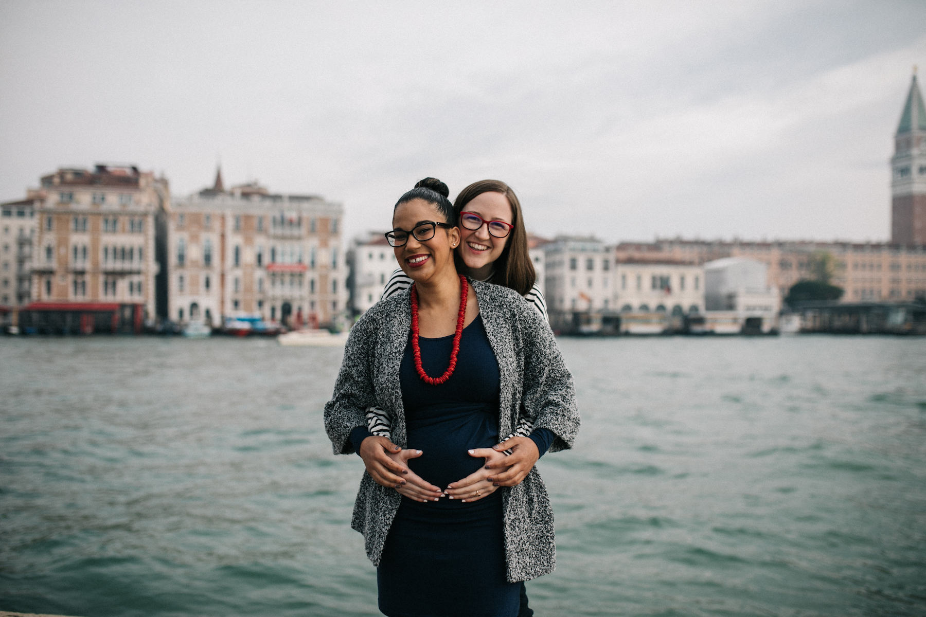 two LGBTQ women holding each other laughing, one of the women is pregnant in Venice, Italy