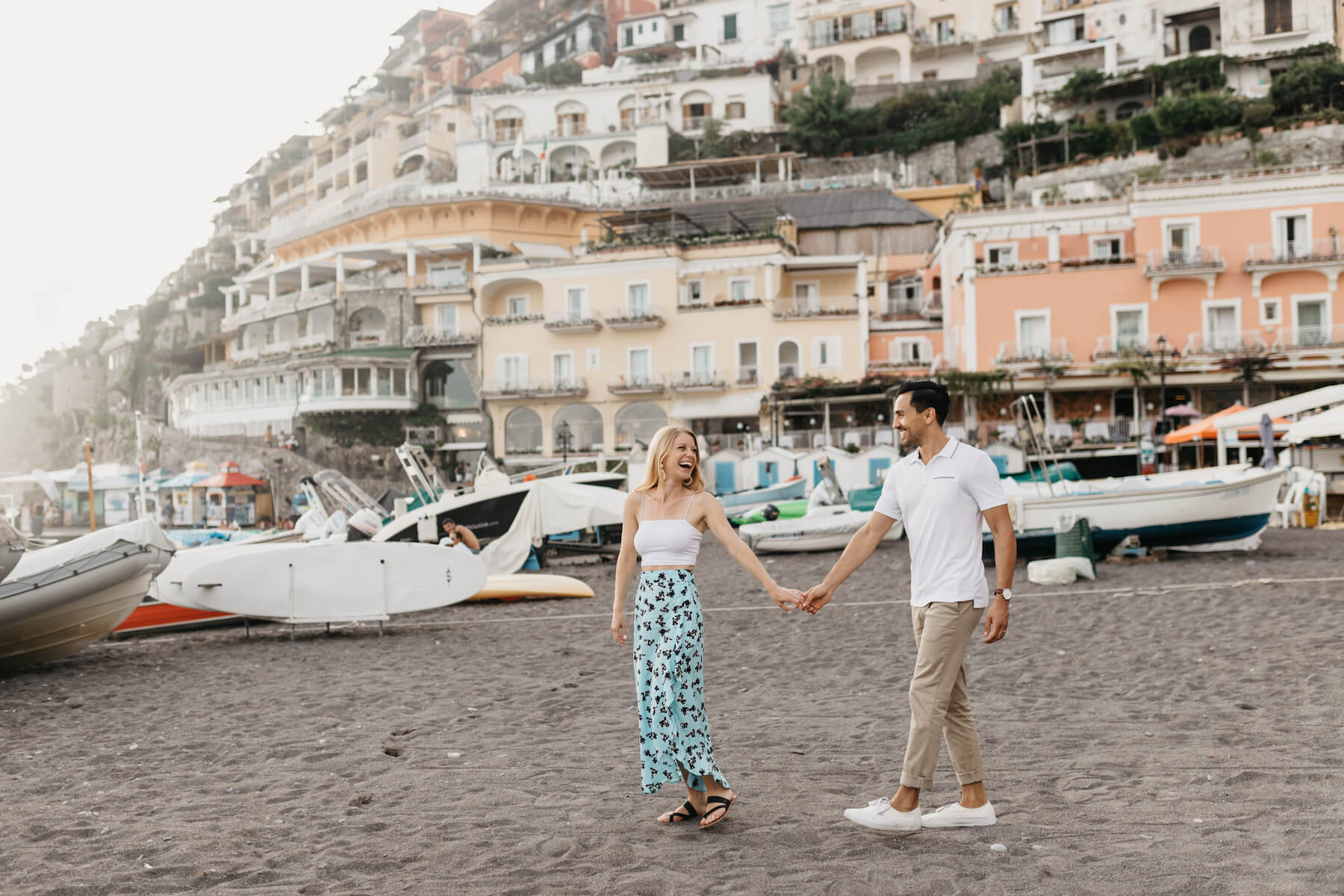 couple holding hands on the beach with the town of Positano behind them, in Positano, Italy
