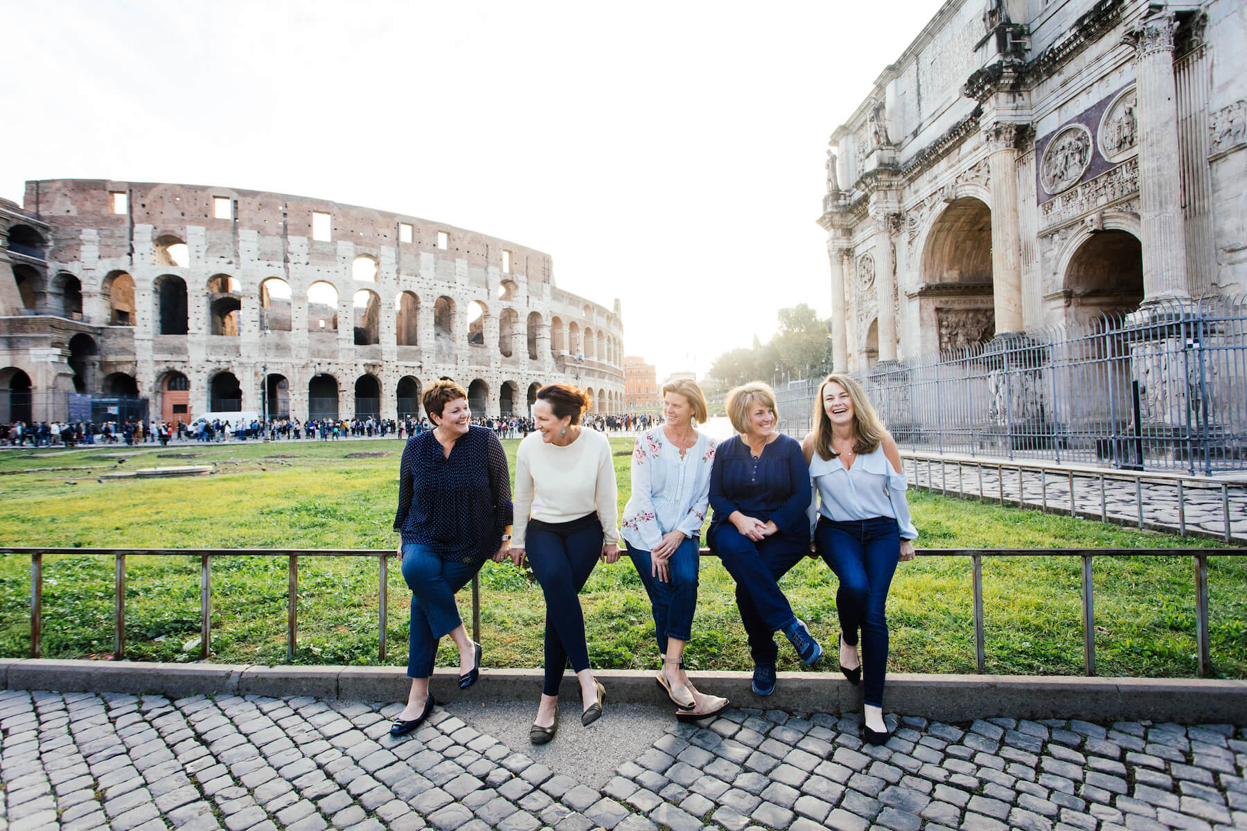 a large group of friends on a girls trip sitting on a fence with the colosseum in the background in Rome, Italy