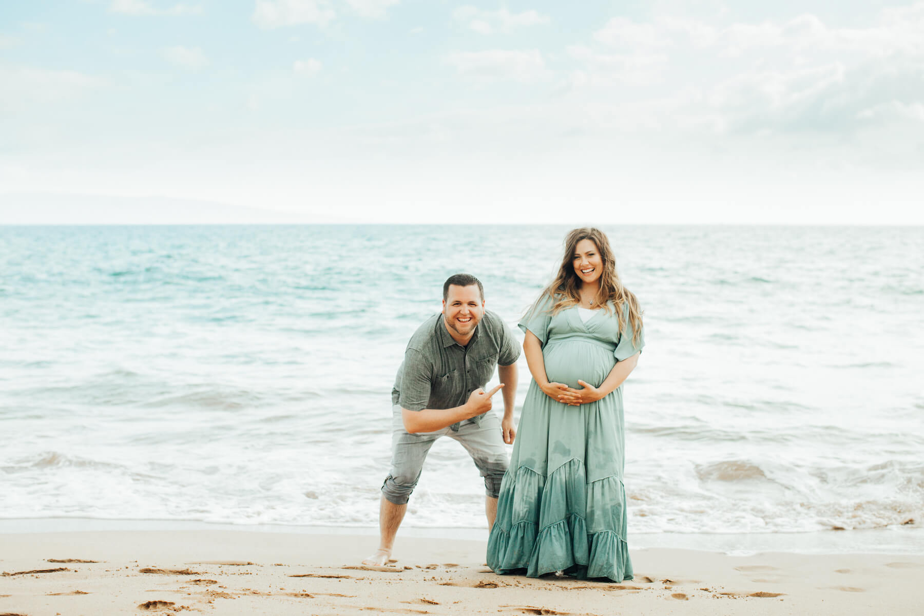 pregnant couple standing on the beach near the ocean and the man is pointing to the woman's belly in Maui, Hawaii