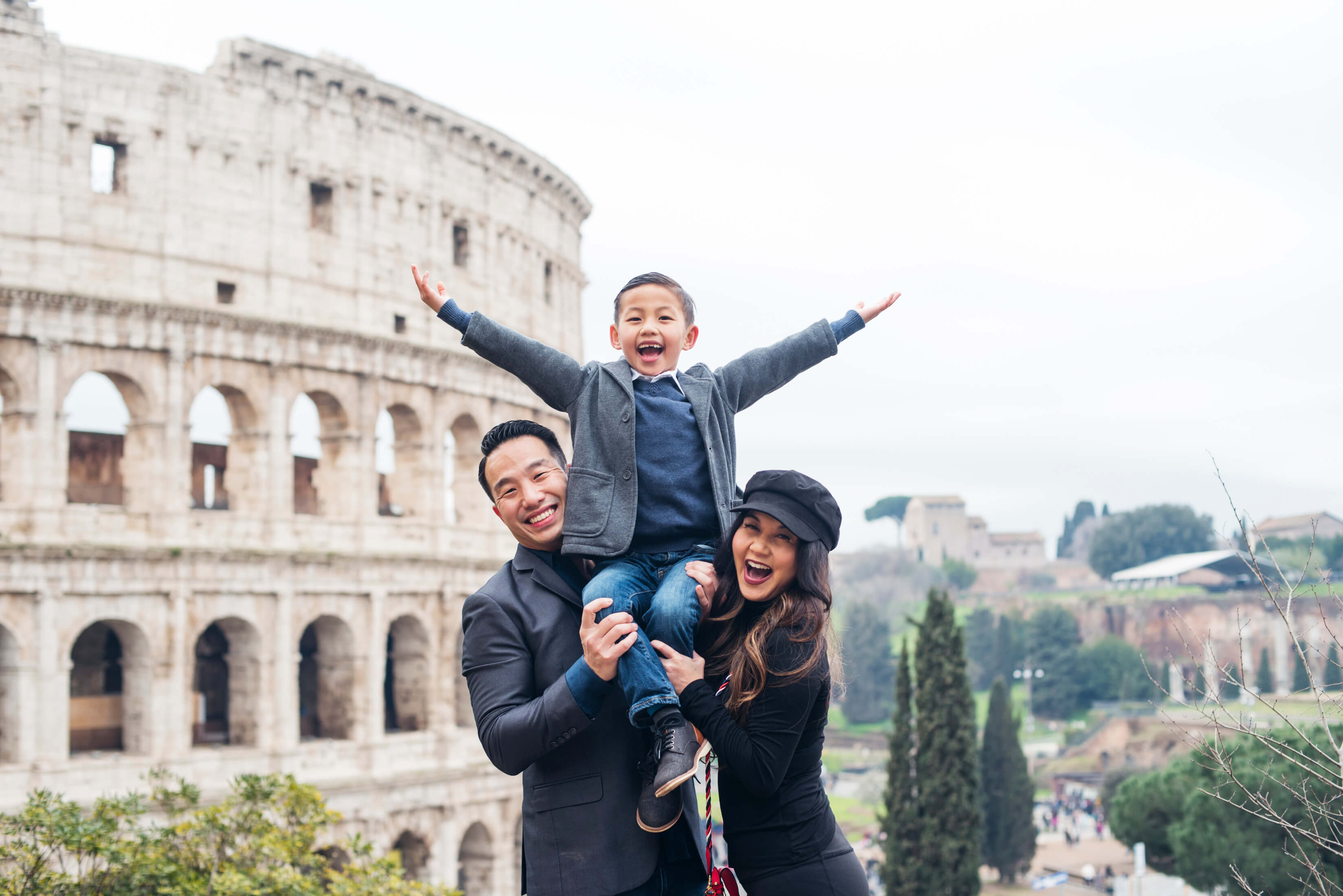 family of three, the son is sitting on his parents shoulders in front of the Roman Colosseum in Rome, Italy