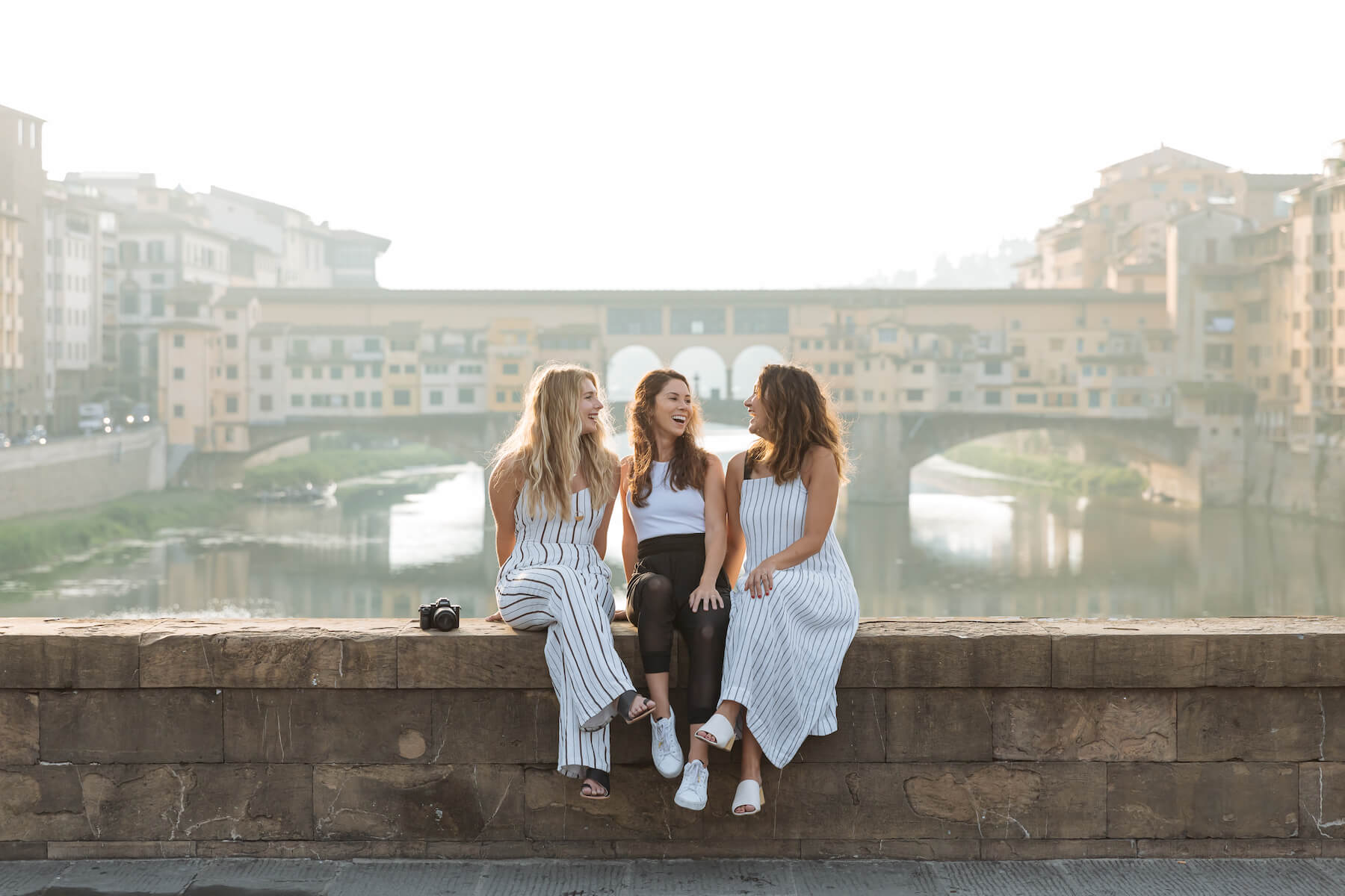 three girlfriends sitting on a bridge ledge with the town of Florence in the background in Florence, Italy