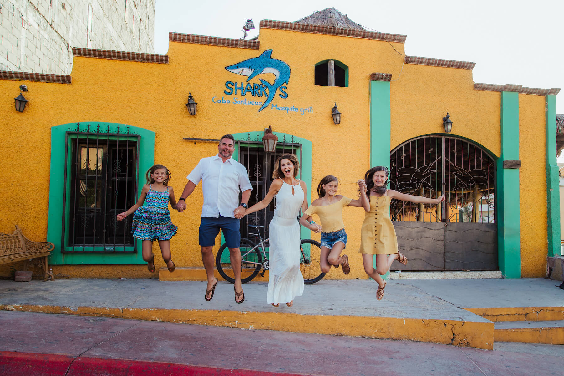 family of five holding hands and jumping in front of a colourful building in Cabo San Lucas, Mexico