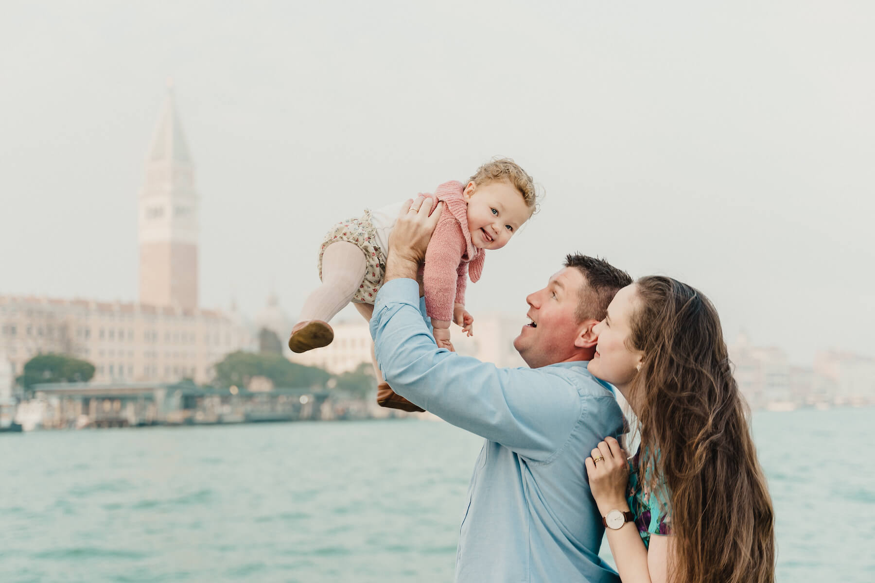 a family of three, the man is holding the baby up in the air with Venice canal in the background in Venice, Italy