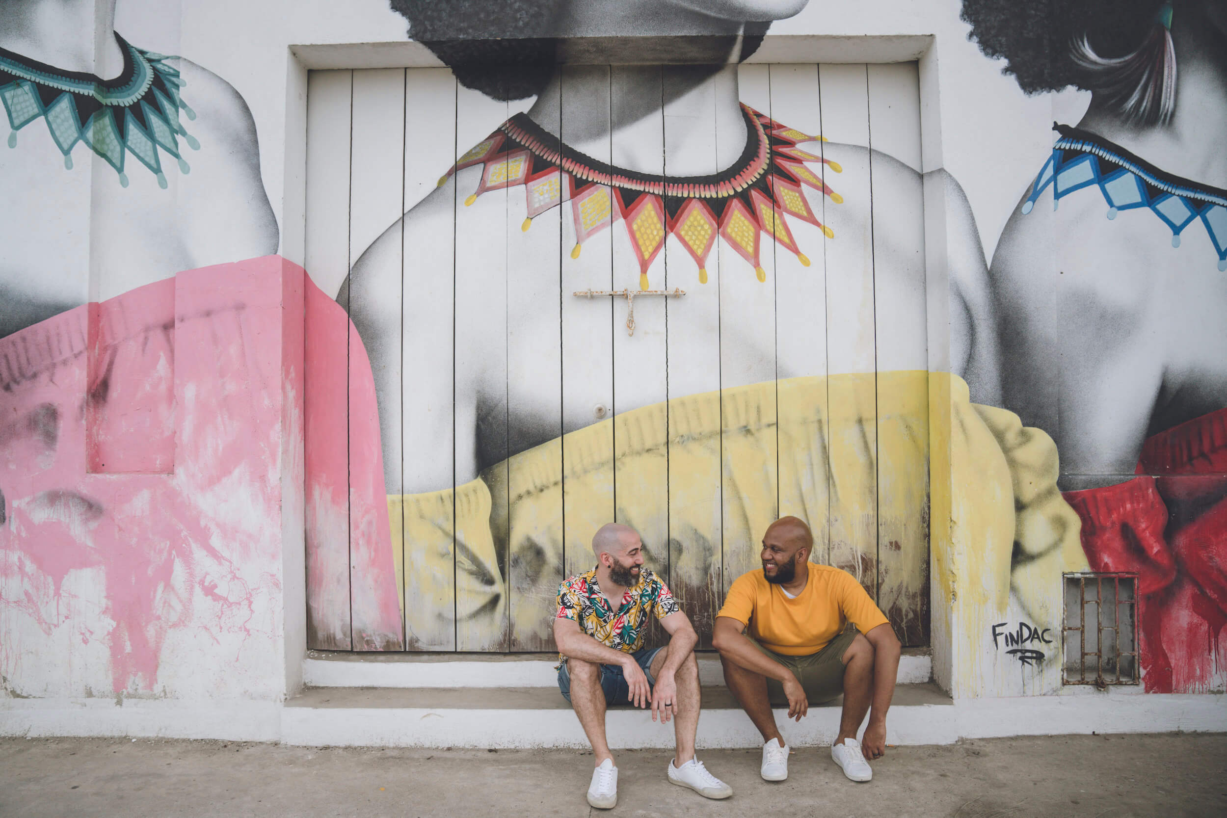 Two men sit on the sidewalk and smile at each other in Cartagena, Colombia.