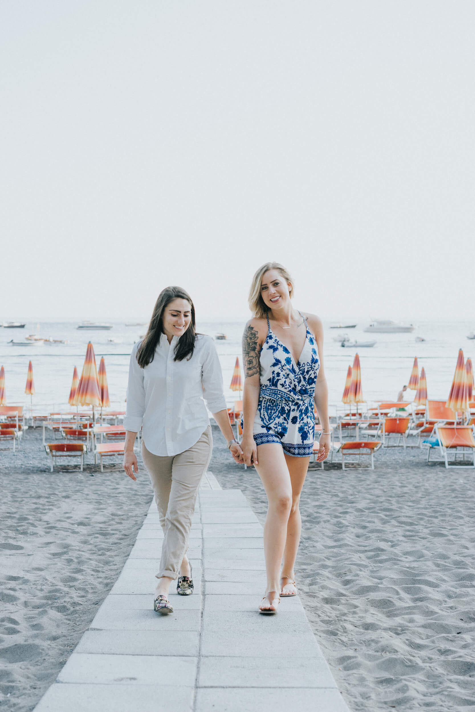 A female couple holds hands as the y walk on the beach in Positano, Italy.