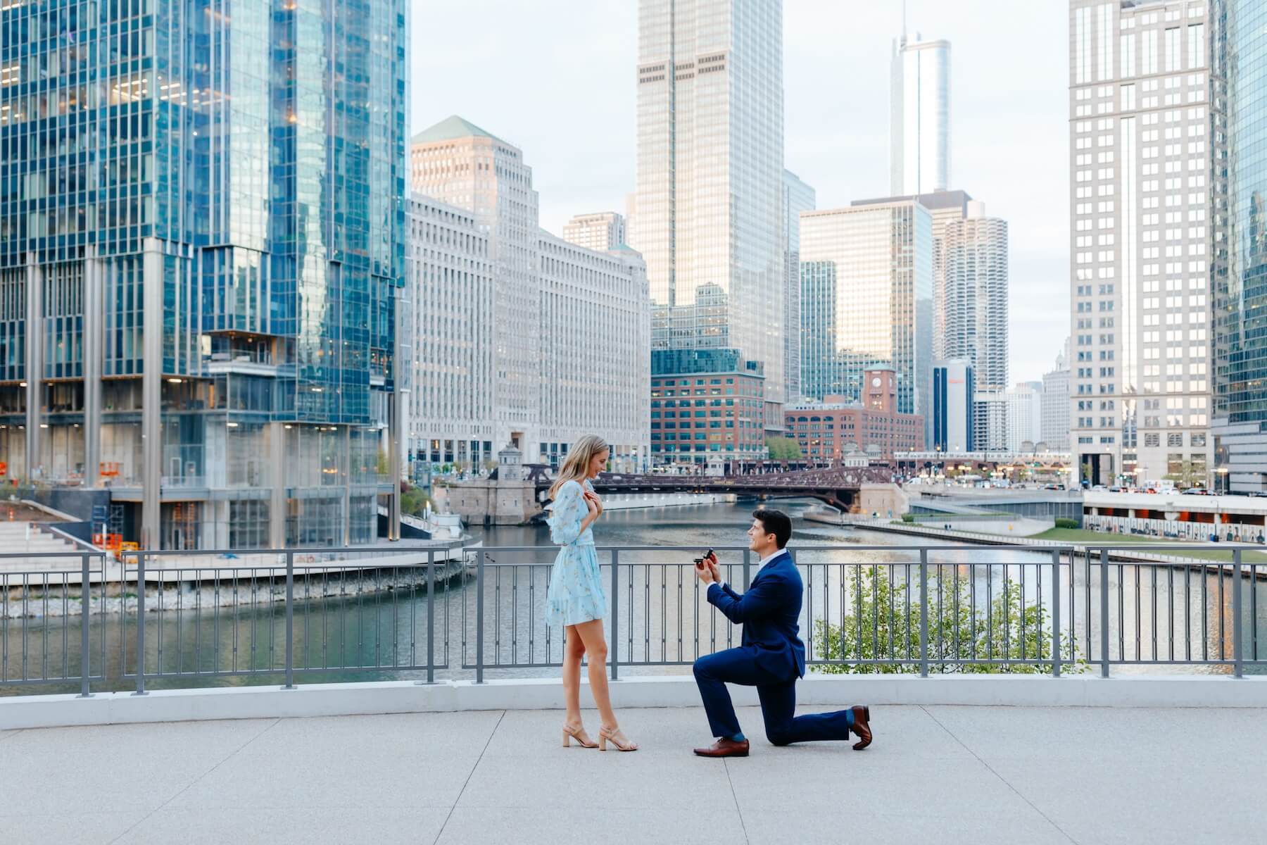 a woman wearing a taupe coloured dress is being proposed to on a bridge in Singapore, the man is wearing a colourful shirt and is on one knee
