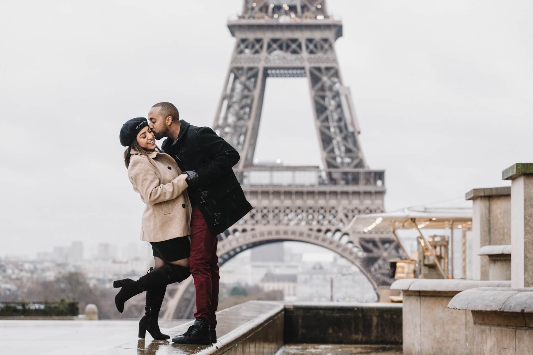 A man kisses his girlfriend on the cheek while they hug in front of the Eiffel Tower.