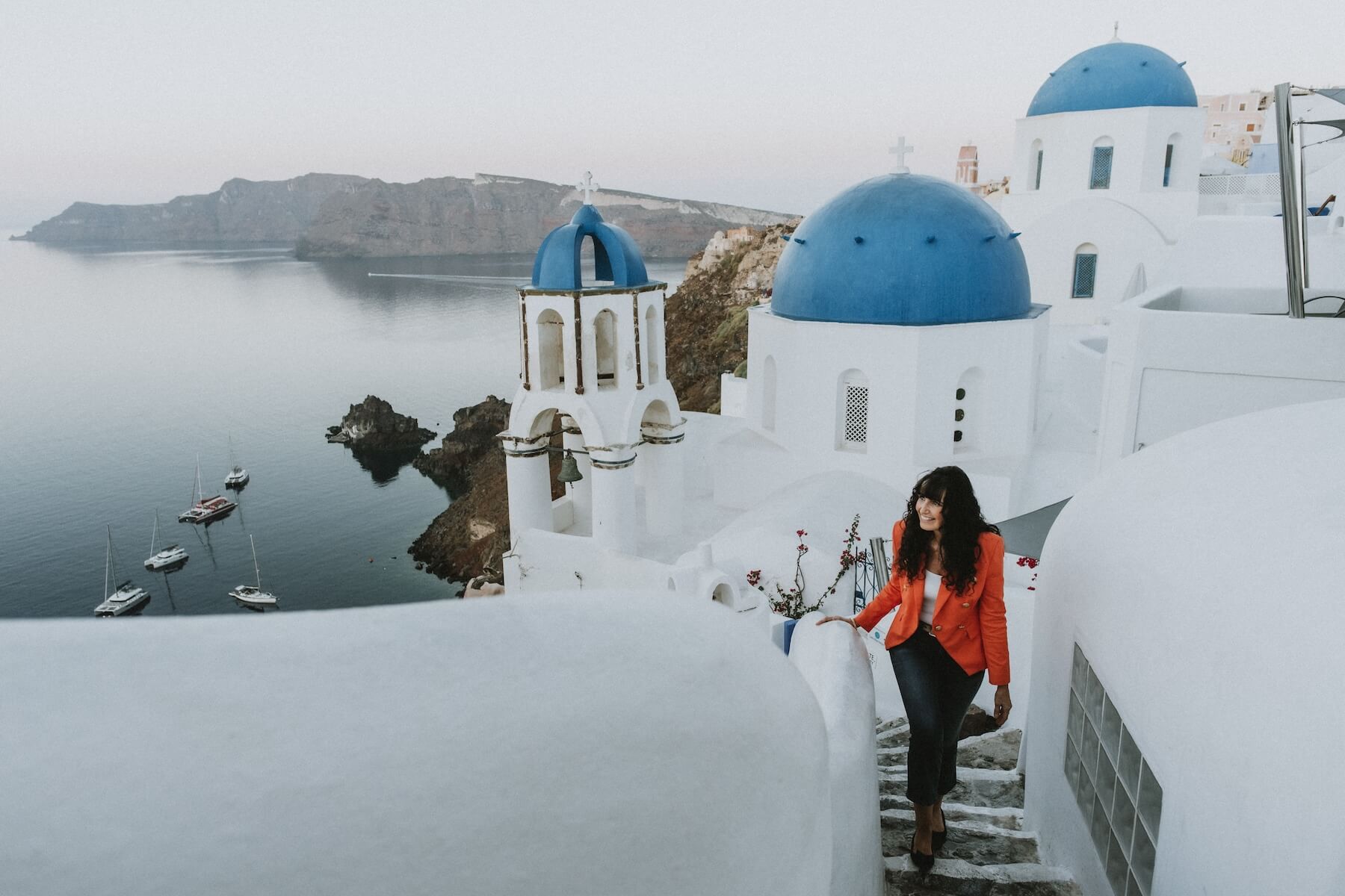 A woman walks up stairs outside with a view of the ocean in Oia Village in Santorini.