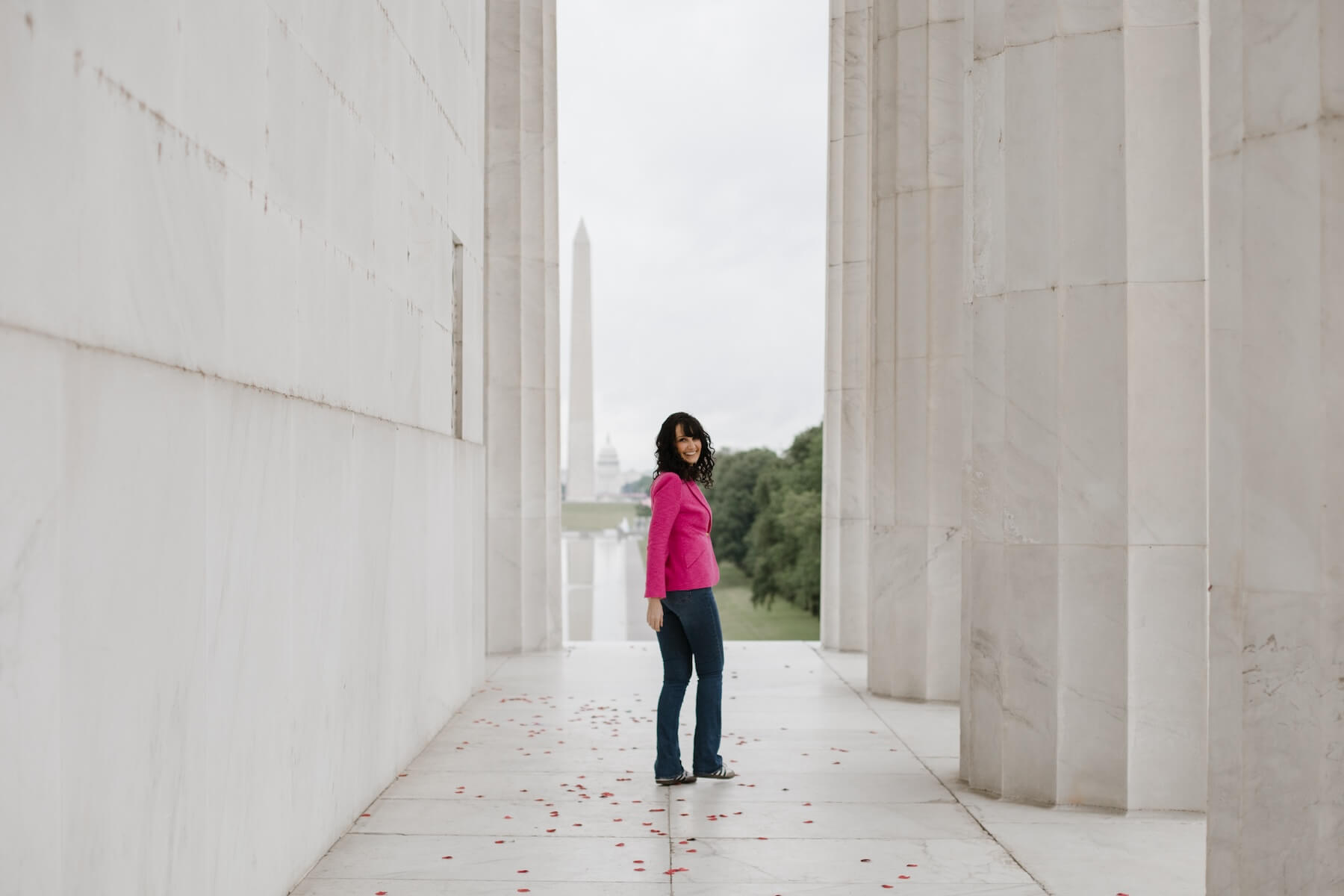 A woman looks back at the camera wearing a pink blazer at the Washington Monument in Washington DC.