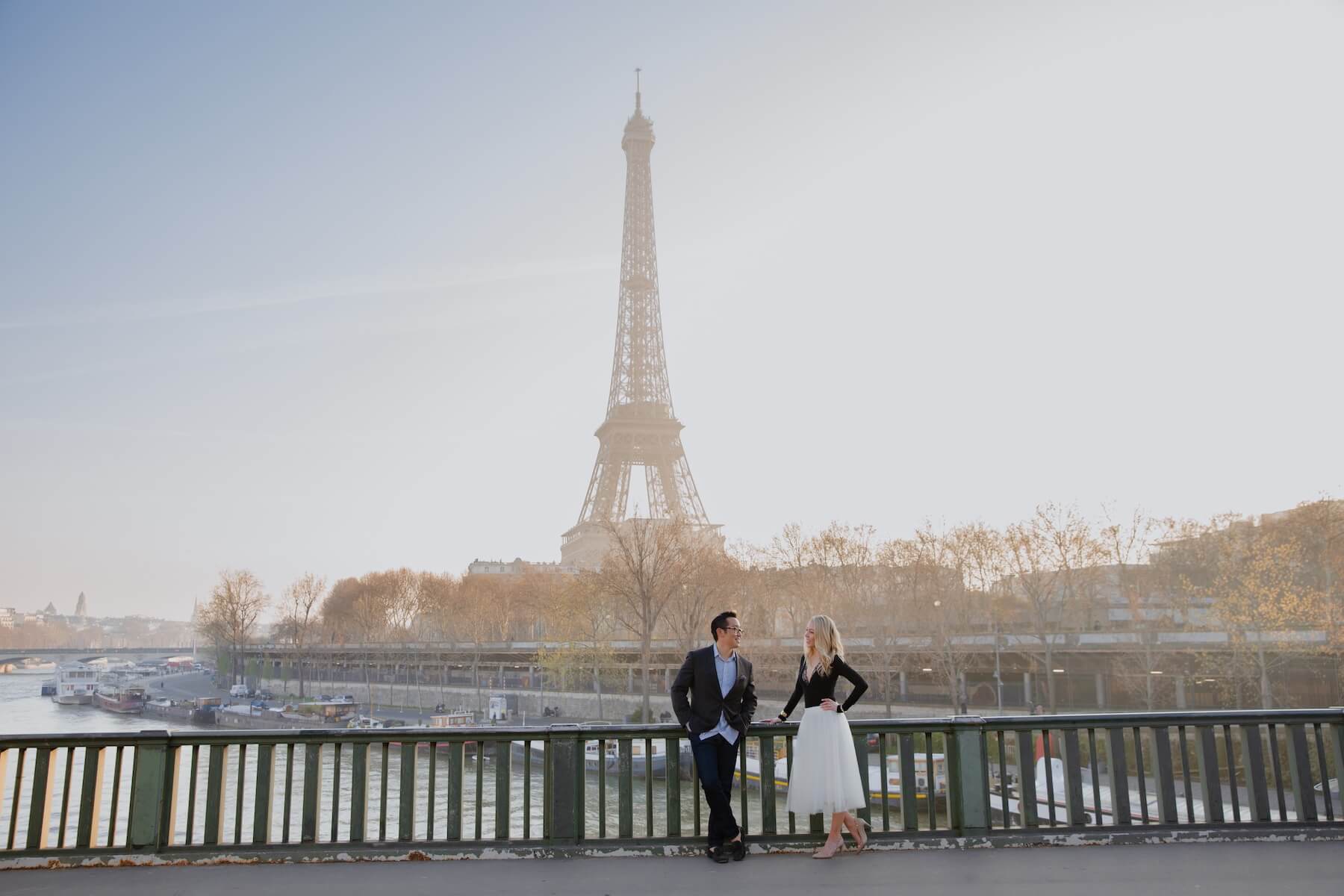A couple look at each other posing at the railing in front of the River and the Eiffel Tower in Paris, France.