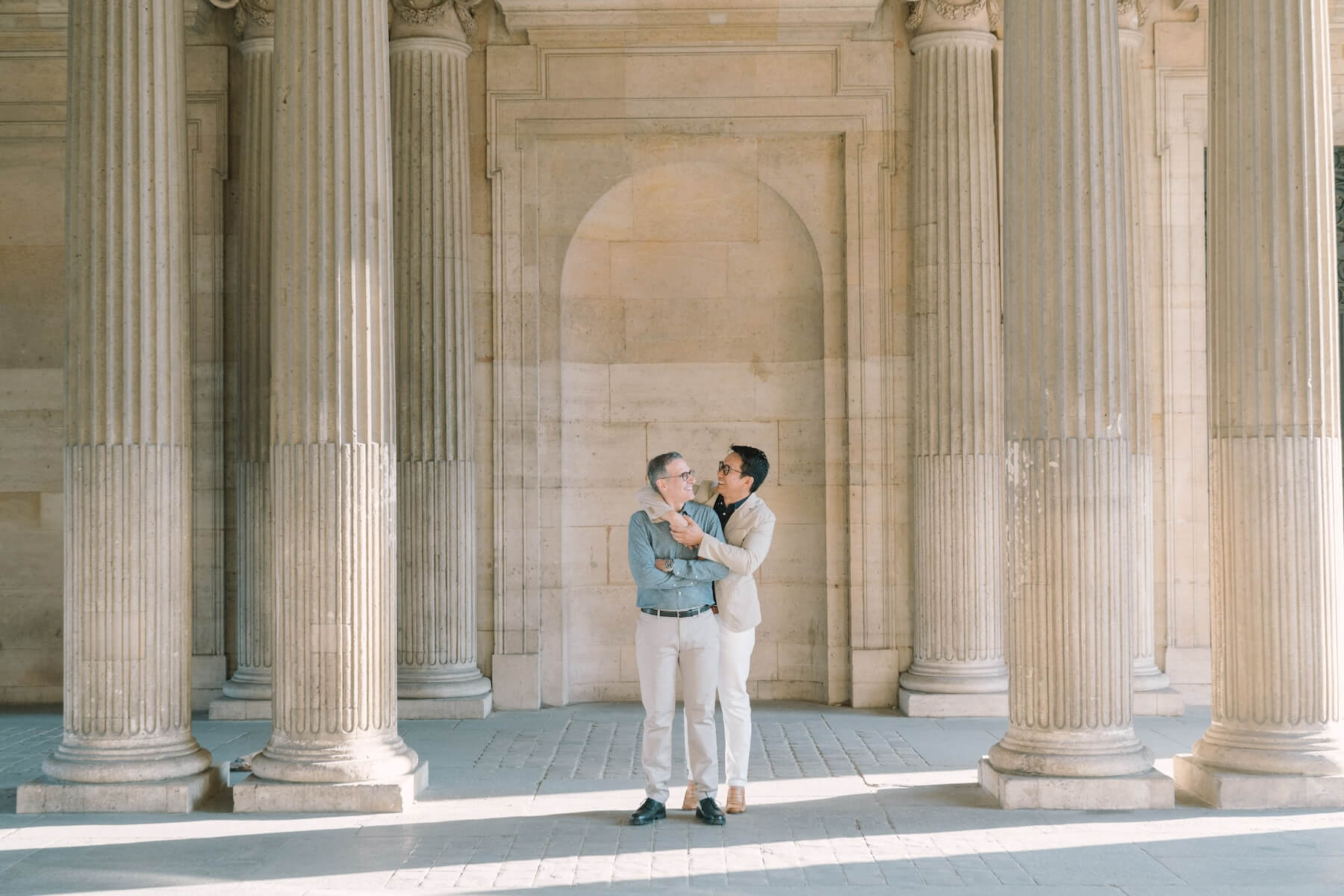 A man wraps his arms around his partner while they smile near the Louvre in Paris, France.