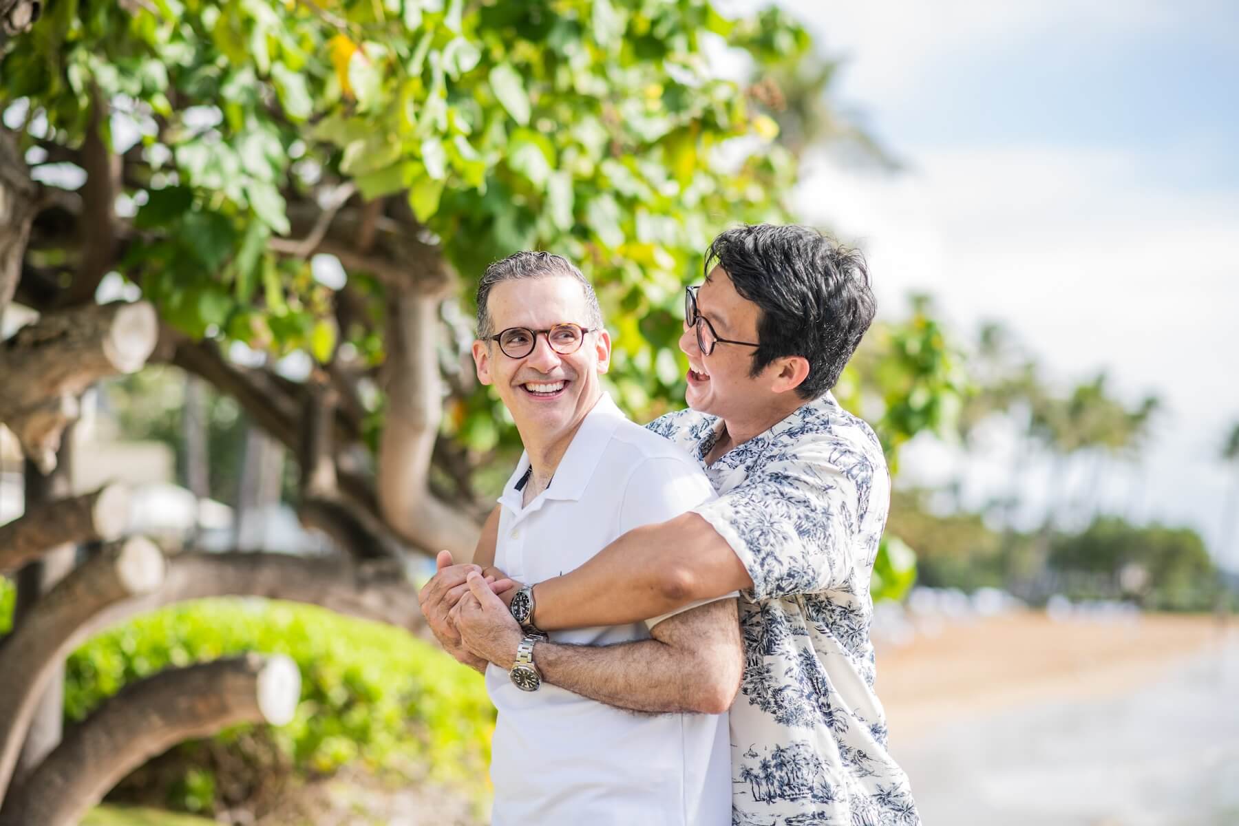 A man wraps his arms around his partner while they smile on the beach in Hawaii.