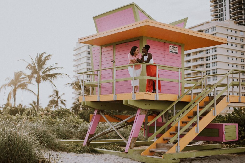 A lesbian couple leaning against a railing of a pink lifeguard station in Miami, Florida at Indian Beach Park on a couples photoshoot with Flytographer.